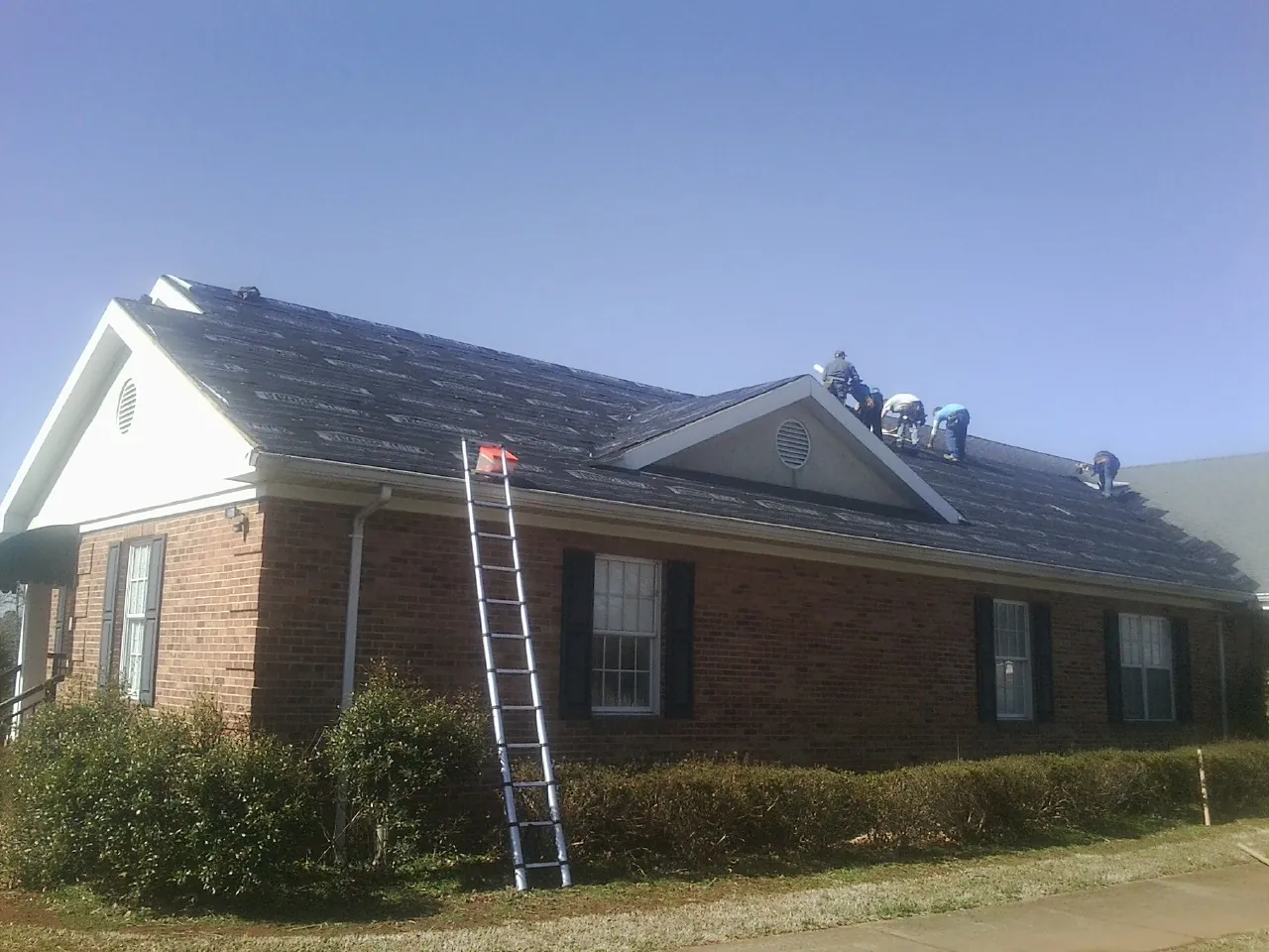 Crew tearing shingles off a brick ranch roof with a ladder set — Metal Roof Repair work by our crew in Haslet