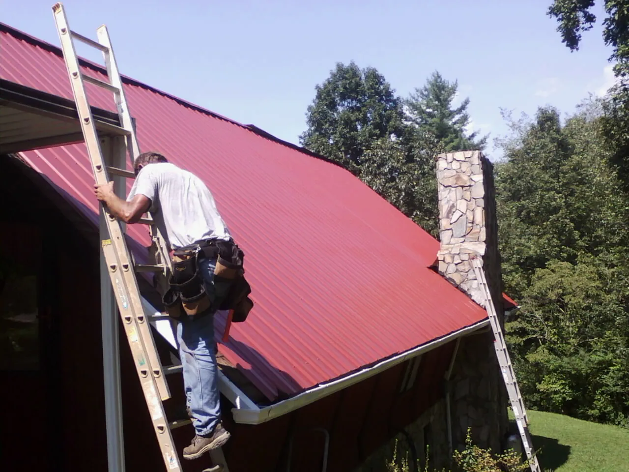 Roofer on a ladder beside a red metal roof and stone chimney — Soffit & Fascia Repair by Roofing Brothers in Haslet